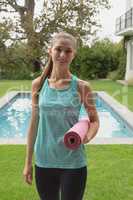 Beautiful female trainer standing with exercise mat in the backyard