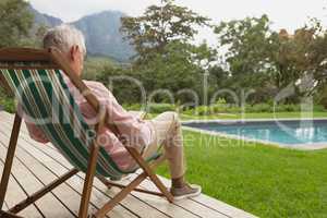Active senior man reading a book while relaxing on sun lounger in the backyard