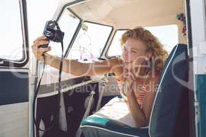 Woman taking selfie with digital camera while lying on seat in camper van at beach