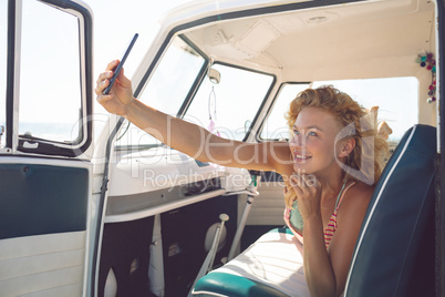 Woman taking selfie with mobile phone while lying on seat in camper van at beach