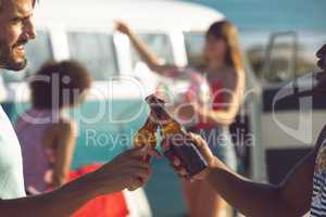 Male friends toasting beer bottle near camper van at beach