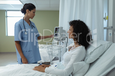 Female doctor adjusting medical monitor while female patient sitting on bed in the ward