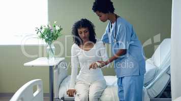 Female doctor helping female patient to get up on hospital bed in the ward