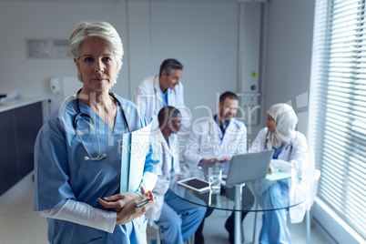 Female surgeon standing with medical file while doctors interacting with each other in the backgroun