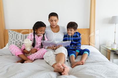 Mother and children reading a story book on bed in bedroom