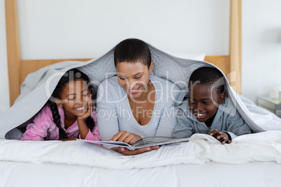 Mother and children reading a story book while relaxing under a blanket in bedroom