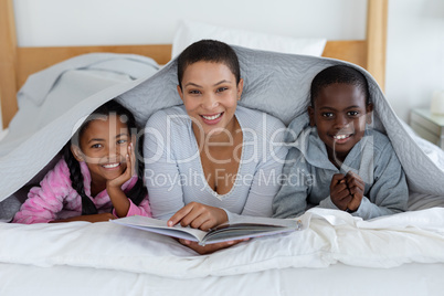 Mother and children reading a story book while relaxing under a blanket in bedroom at home