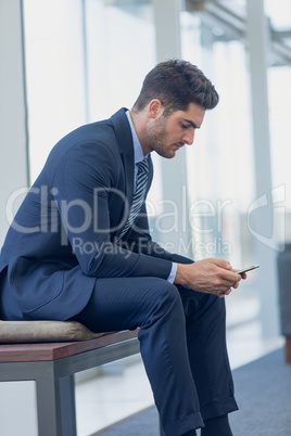 Caucasian businessman looking at mobile phone while sitting on bench in modern office.