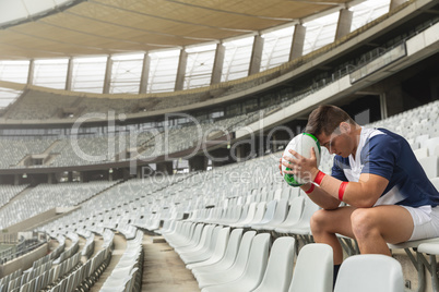 Upset Caucasian Male rugby player sitting with rugby ball in stadium