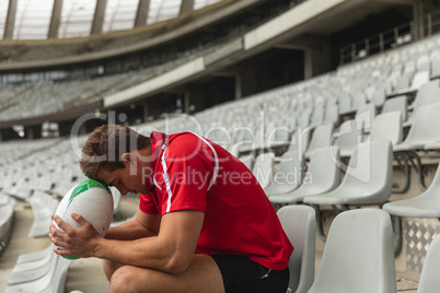 Upset Caucasian male rugby player sitting with rugby ball in stadium