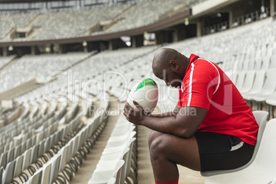 Upset African American male rugby player sitting with rugby ball in stadium