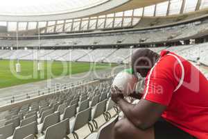 Upset African American male rugby player sitting with rugby ball in stadium