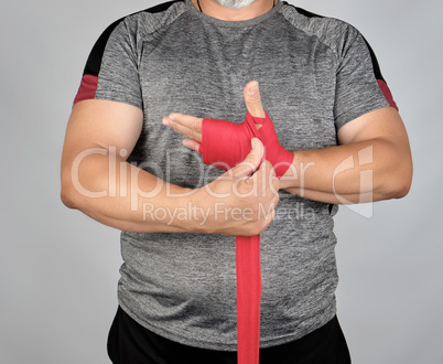 athlete stand in gray clothes and wrap his hands in red textile