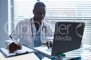 Male doctor writing on a clipboard while using laptop at desk