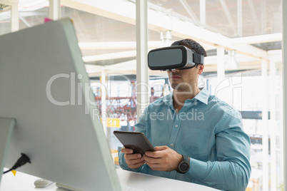 Male executive holding digital tablet while using virtual reality headset at desk