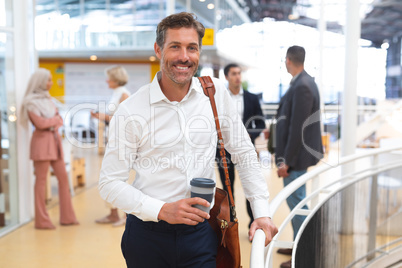 Businessman holding disposable coffee cup and looking at camera near railing in a modern office