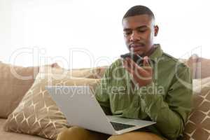 Man talking on mobile phone while using laptop on sofa in living room at comfortable home