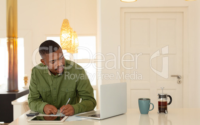 Man looking at laptop screen and writing on paper at dining table in kitchen at comfortable home