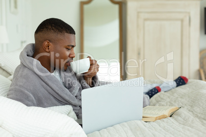 Man drinking coffee while using laptop on bed in bedroom