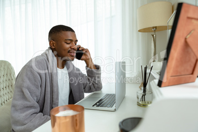 Man talking on mobile phone while working on laptop at desk in bedroom