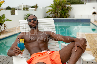 Man in sunglasses sitting with cocktail glass on sun lounger near swimming pool