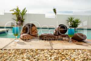 Couple with cocktail glasses leaning on the edge of a swimming pool