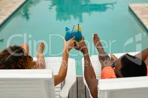 Couple toasting glasses of cocktail while relaxing on a sun lounger near swimming pool at the backya
