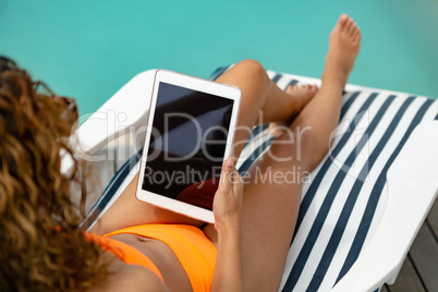 Woman using digital tablet while relaxing on a sun lounger near swimming pool at the backyard of hom