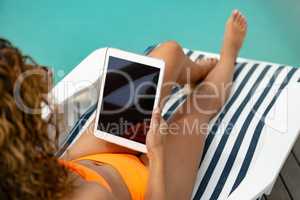 Woman using digital tablet while relaxing on a sun lounger near swimming pool at the backyard of hom