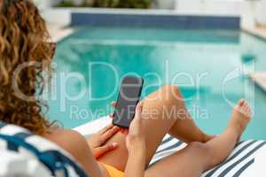 Woman using mobile phone while relaxing on a sun lounger near swimming pool at the backyard of home