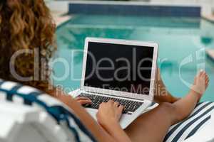 Woman using laptop while relaxing on a sun lounger near swimming pool at the backyard of home