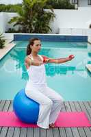 Woman exercising with resistance band while sitting on a exercise ball near swimming pool in the bac