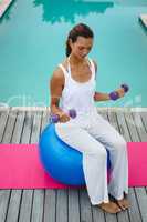 Woman exercising with dumbbells while sitting on a exercise ball near swimming pool in the backyard