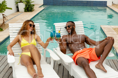 Couple toasting glasses of cocktail while relaxing on a sun lounger near swimming pool