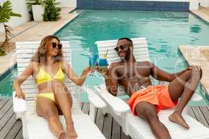 Couple toasting glasses of cocktail while relaxing on a sun lounger near swimming pool