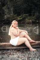 Junge kurvige Frau sitzt wenig bekleidet an einem See auf einem Baumstamm