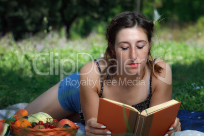 Girl lying in the park grass, reads a book and eats fruit.