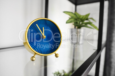 Popular round clock with hour and minute hands on glossy surface in office. Small golden tabletop clock with blue analog face on glass shelf. Decorative mantel clock on rack at home. Quartz movement