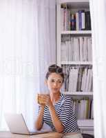 Taking a break from her blogging. Shot of a young woman sitting with a laptop and holding a beverage.