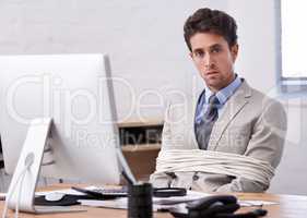 Hes always stuck to his desk. A handsome young businessman tied up and sitting at his desk.