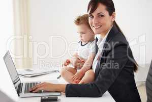 Managing career and motherhood with ease. Young working mom smiling at the camera while holding a camera and typing on her laptop.