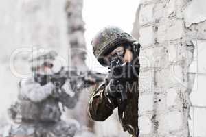 Dynamic defense force. A soldier peering around a wall while pointing a gun at the camera during a rade on a damaged building.