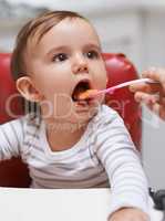 Open wide.... Shot of a cute little baby sitting in a high chair and being fed by her mom.