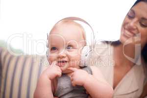 What you listening to. Shot of an adorable baby girl wearing headphones while sitting with her mother.
