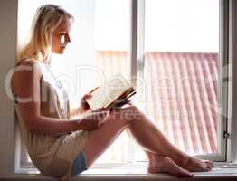 Engrossed in her novel. A young woman relaxing on a window sill and reading an absorbing novel.