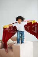 Flying around with his imagination. A cute young boy pretend to fly his lounge sofa holding a blanket like a superheros cape.