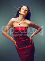 Teasing temptress. Fashion model posing sensually in a fitting red satin dress against a dark background.