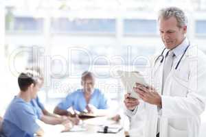 Doing his daily check. A senior doctor holding a touchpad with colleagues sitting in the background.