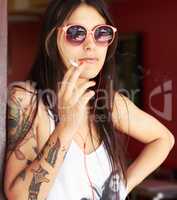 Rebel at heart. Shot of a beautiful young tattooed woman smoking a cigarette while listening to music.