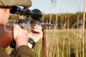 Patience is the secret to a perfect shot. A game hunter looking through the reeds with his sniper rifle.